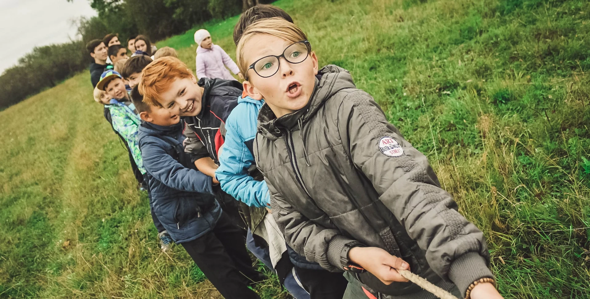 Children participating in a tug of war in a grassy field during the Youth Initiative event, with one boy in the foreground wearing glasses and displaying a surprised expression.
