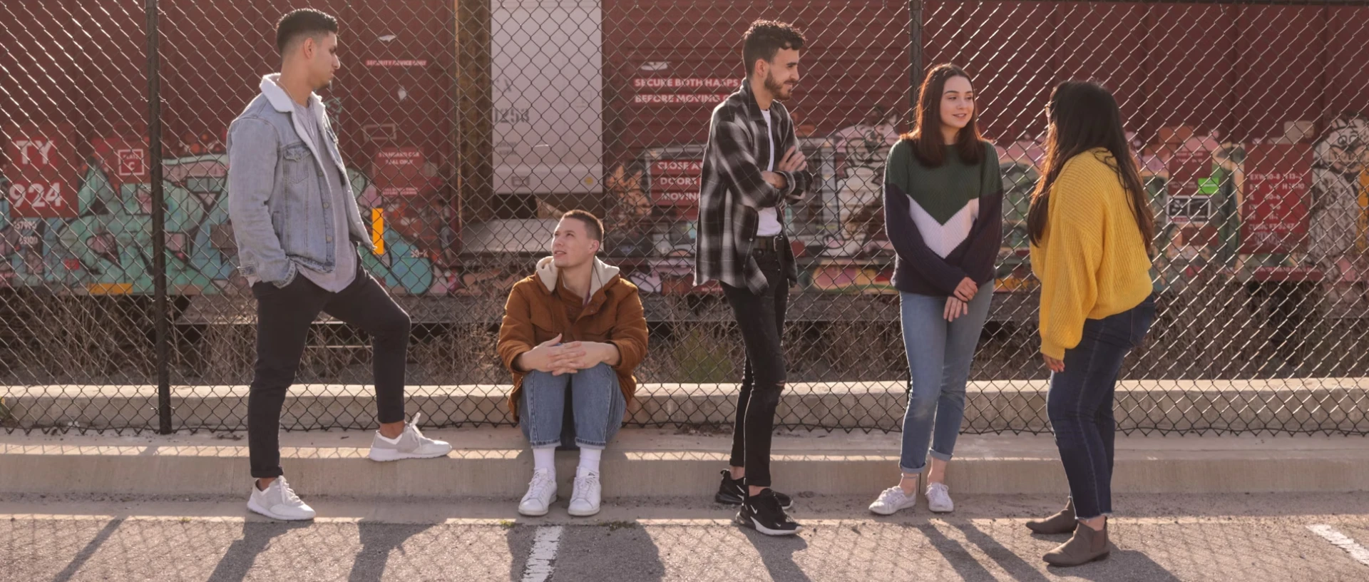 Five young adults chatting casually in front of a graffiti-covered wall, with one sitting on a short fence, participating in the Youth Initiative program.