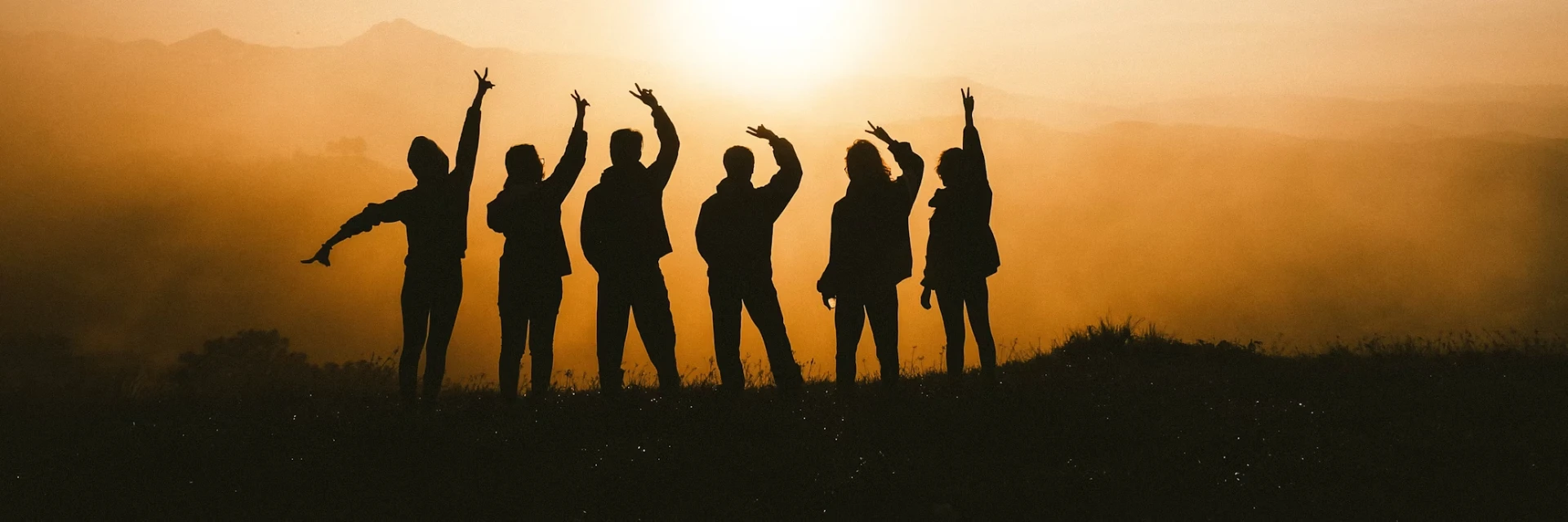Silhouettes of seven people with raised arms standing on a hill, backlit by a golden sunset with hazy mountains in the background, celebrating the visit of Her Excellency Sharon Johnson.