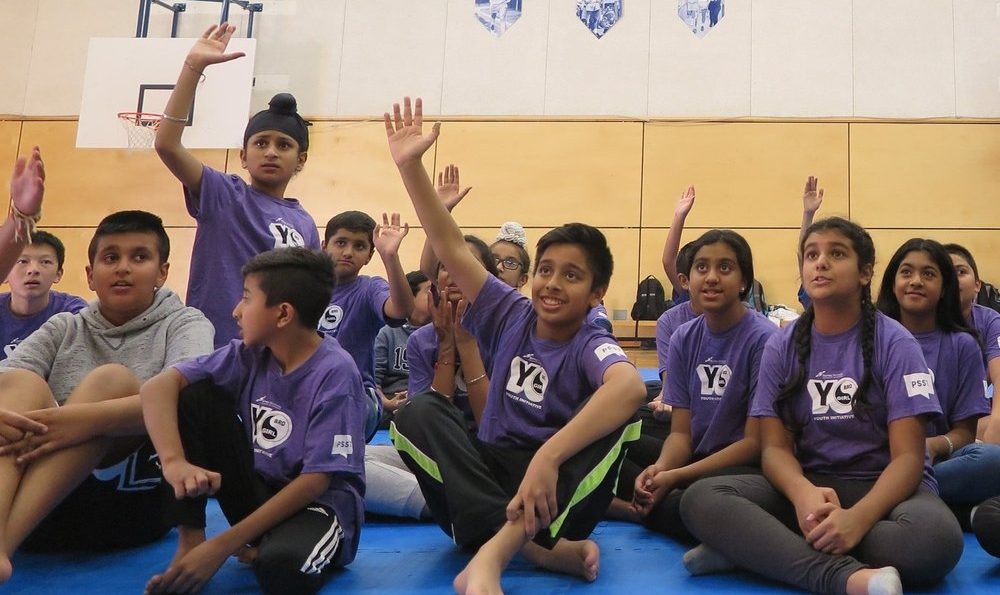 Children in purple shirts sitting on a gym floor, raising their hands and looking attentively forward during a group activity. For further details, contact us.