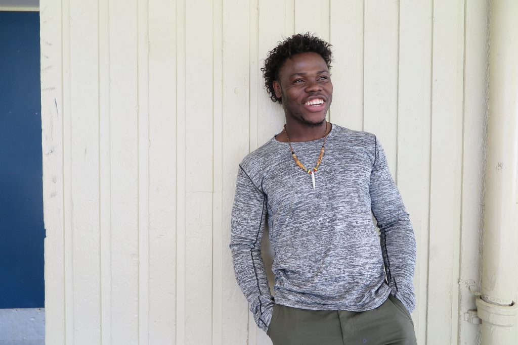 A young man with curly hair, laughing and standing against a cream paneled wall, wearing a gray long-sleeved shirt and olive pants.