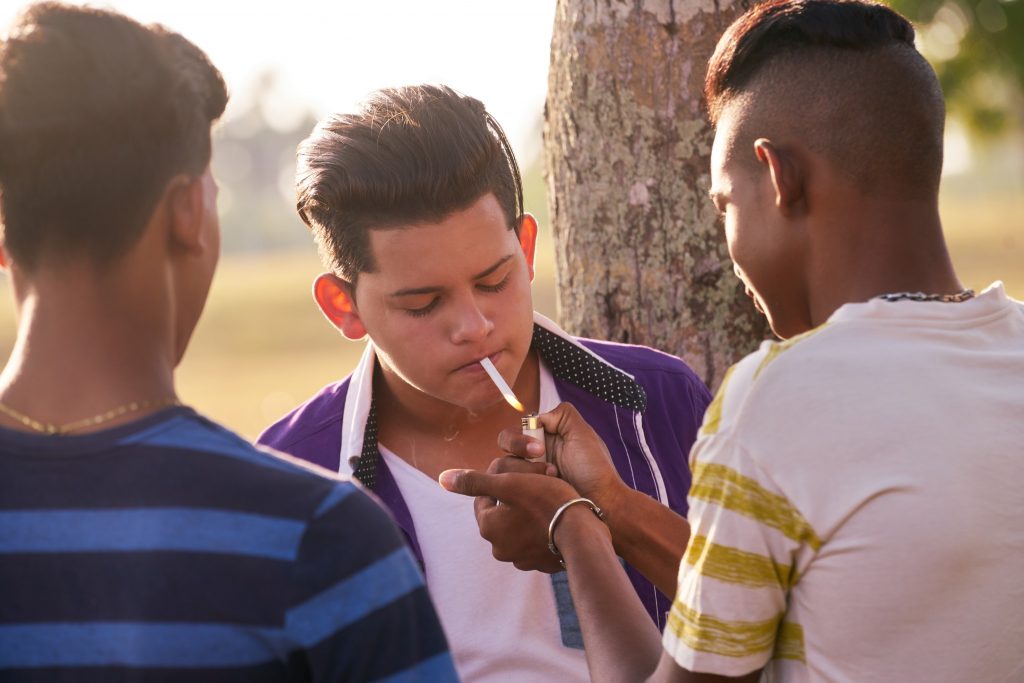 A young man lighting a cigarette while flanked by two friends outdoors, with a warm sunset background, evoking thoughts of youth gangs.