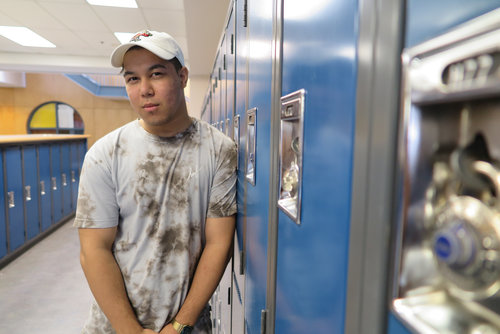 Young man in a tie-dye shirt standing in front of blue lockers in a school hallway, holding educational resources.