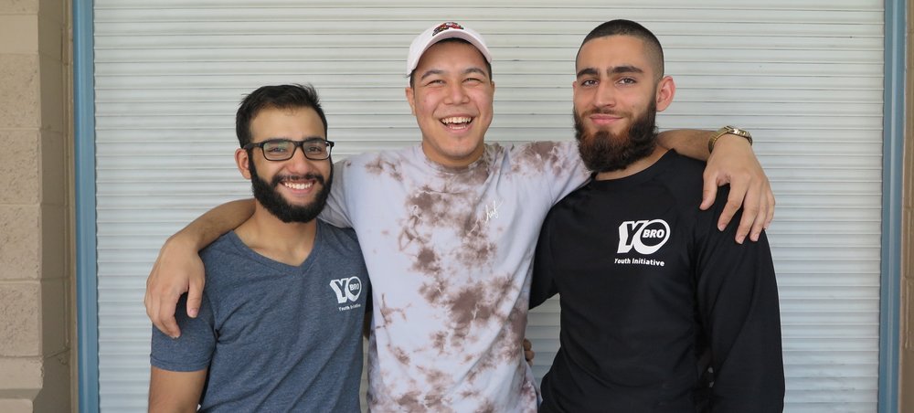 Three young men smiling and posing arm-in-arm in front of a metal garage door. One wears glasses, another a cap, and the third a black shirt with a logo. For better service, it