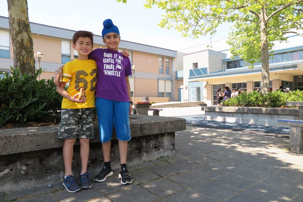 Two boys smiling and standing together in a schoolyard on a sunny day; one wearing a yellow shirt and camo shorts, the other in a purple shirt and blue jeans.