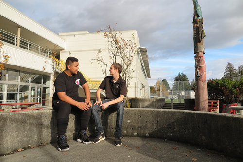 Two young men sitting and talking on a concrete ledge outside a modern building with resources like a totem pole in the background.