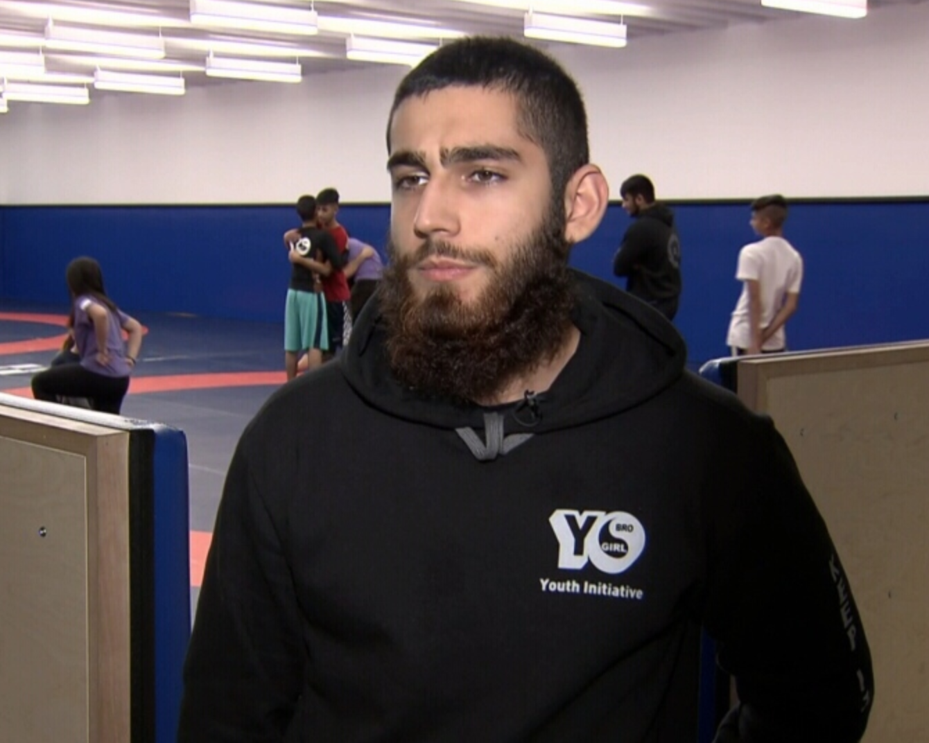 A young man with a beard wearing a black hoodie with a "youth initiative" logo, representing a local group, sits indoors while kids from gangs practice martial arts in the background.